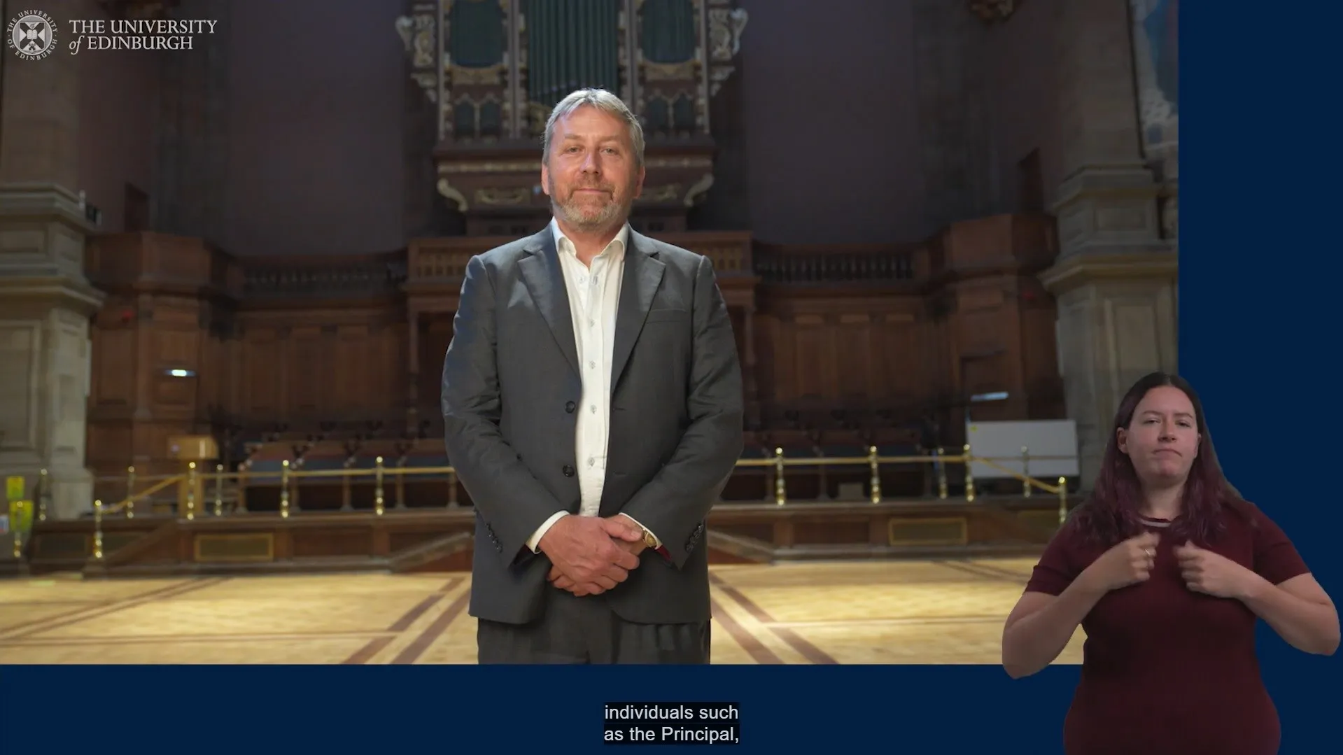 Screenshot from the Principal's Welcome Ceremony with Prof Peter Mathieson in the centre of the picture alongside the BSL translator in the bottom right corner. The Principal is smiling at the camera as he stands in McEwan Hall and at the bottom of the picture there are captions which read: 'individuals such as the Principal'